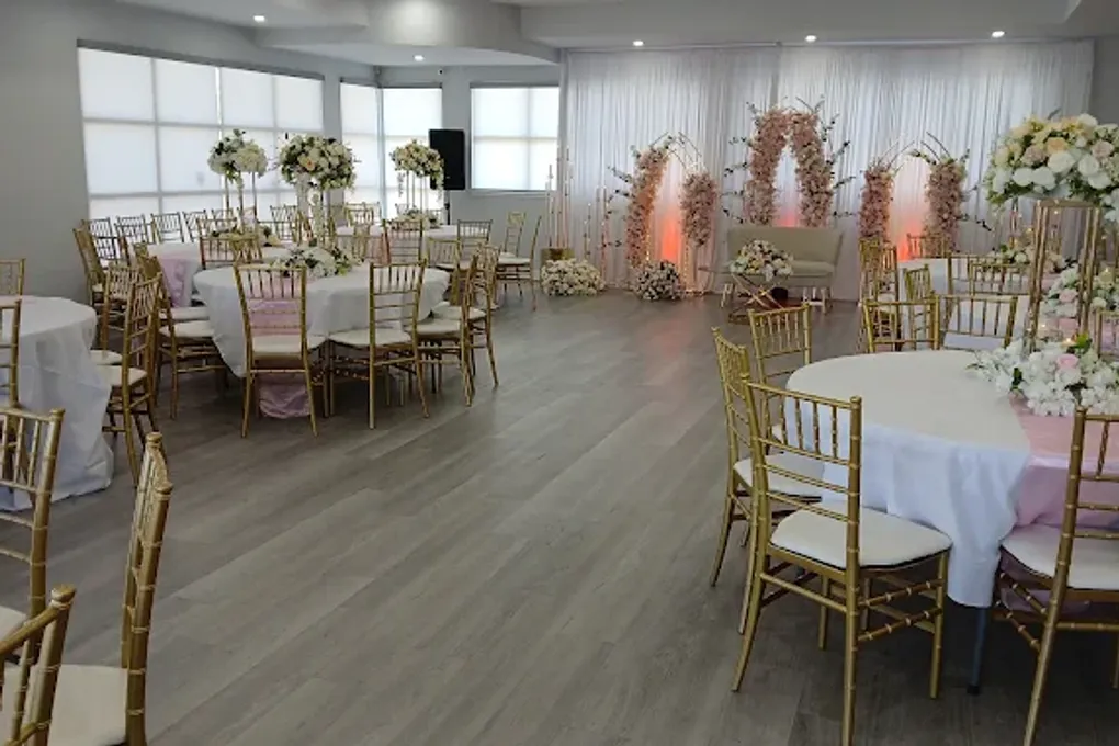 The banquet hall laid for a wedding: round tables in white and blush, gold chiavari chairs, and a floral arch against draped walls.