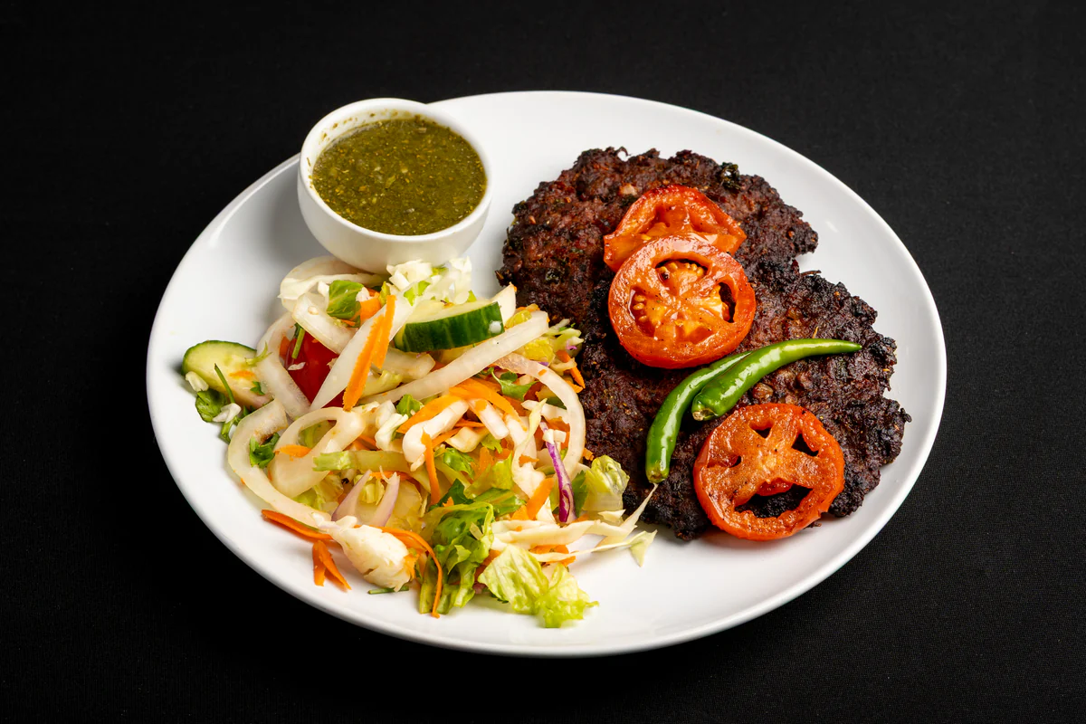 A chapli kabob on a white plate, topped with grilled tomato and green chillies, shredded salad and green chutney alongside.