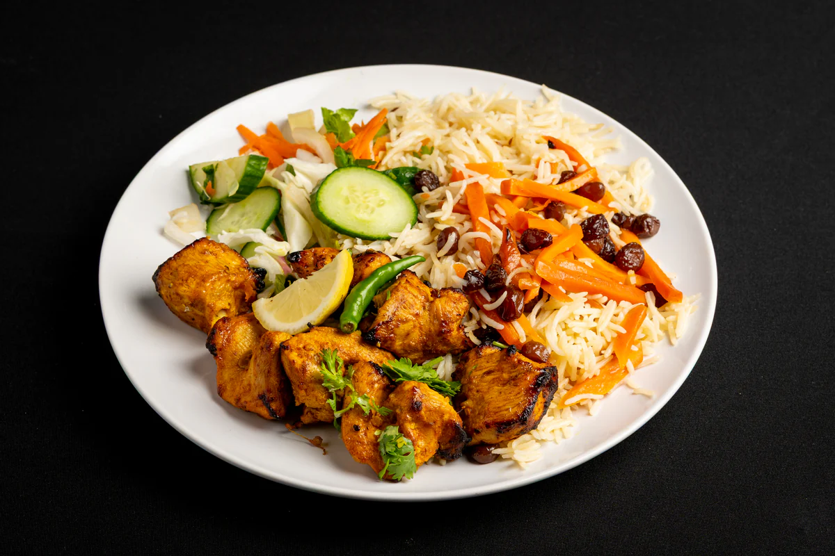 Turmeric-marinated chicken tikka, charred at the edges, beside pulao rice with carrot and raisins, salad and a lemon wedge.