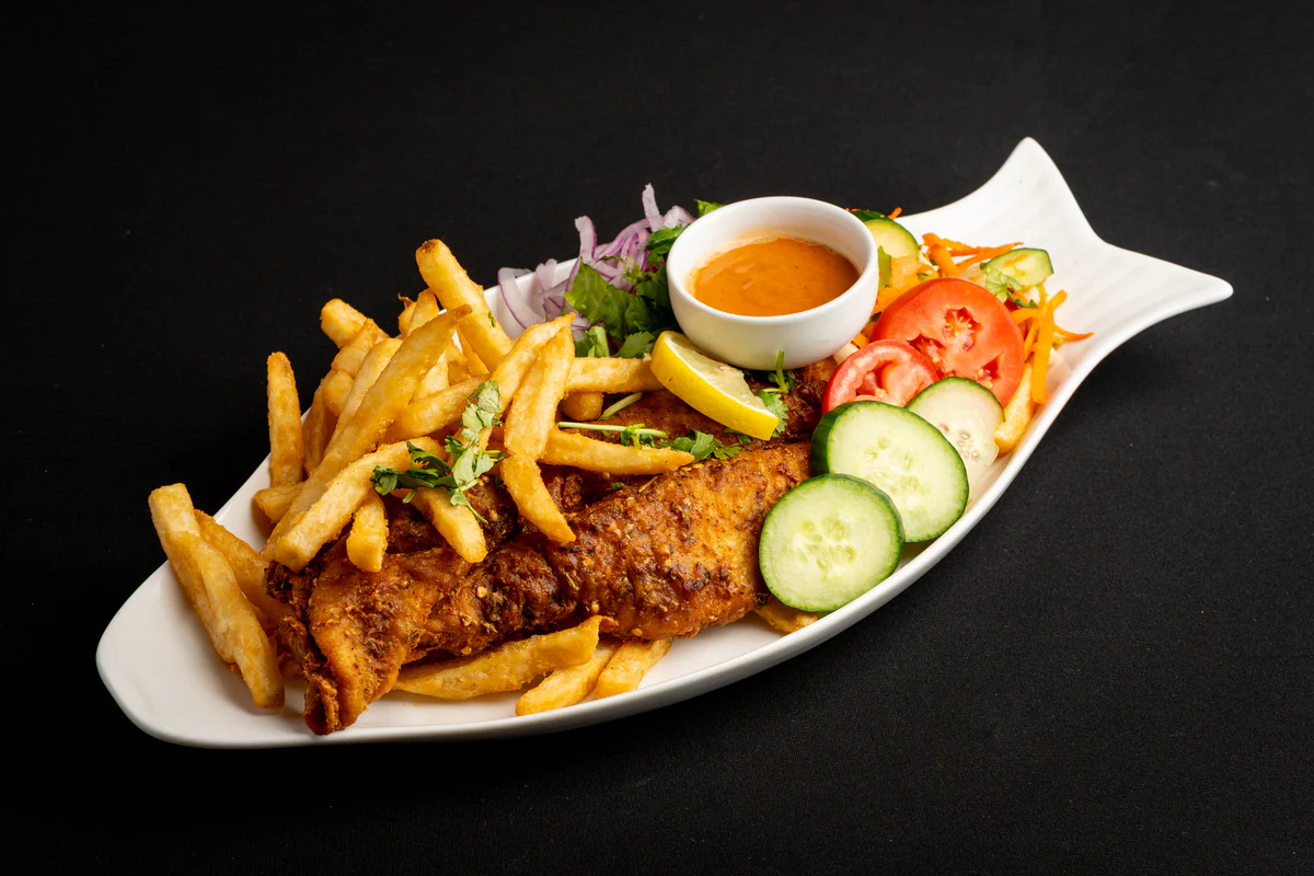 A fried fish fillet in a crisp spiced crust with french fries, on a curved white plate with lemon, salad and a dipping sauce.