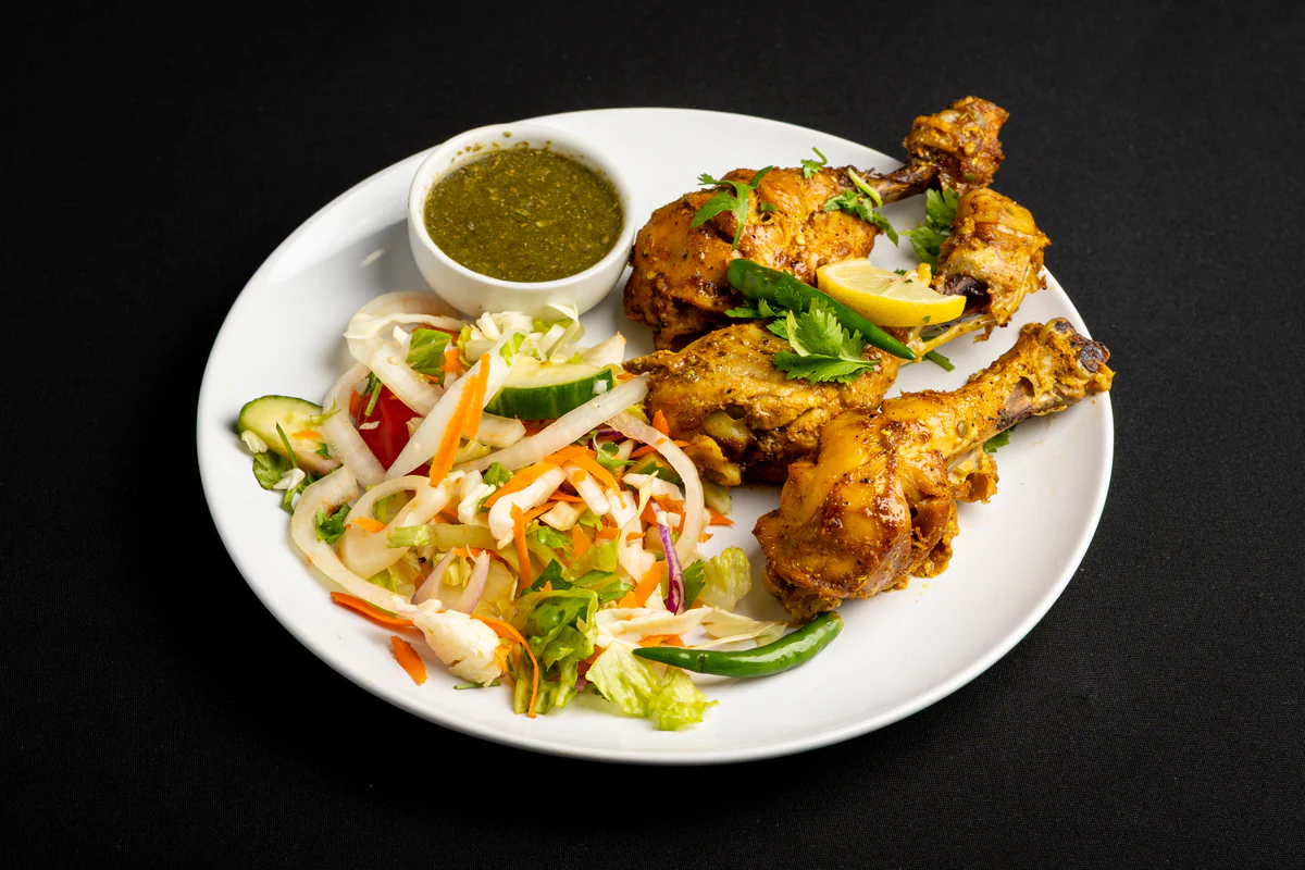 Grilled chicken on the bone, golden with spice, on a white plate with green chutney, shredded salad and a green chilli.