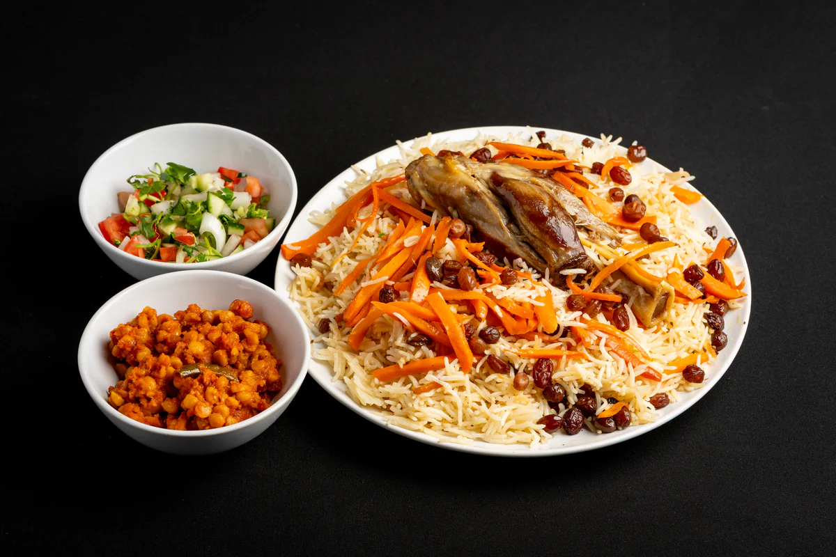 A whole braised lamb shank set into a plate of Kabuli pulao, the rice ridged with caramelised carrot and raisins, with chana and chopped salad alongside.