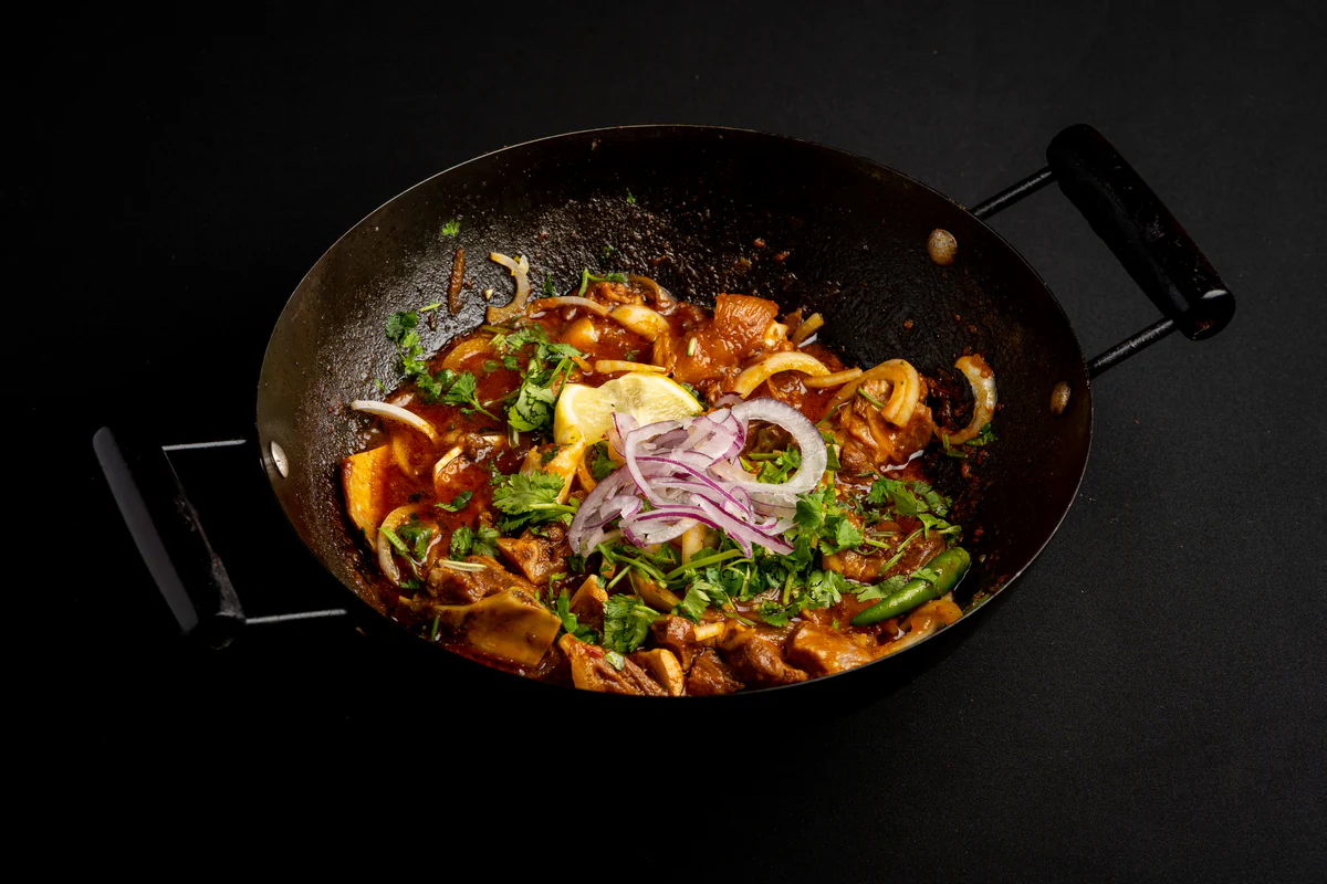 Lamb karahi in a black wok, bone-in pieces in a red sauce under coriander, sliced onion, a lemon wedge and a green chilli.