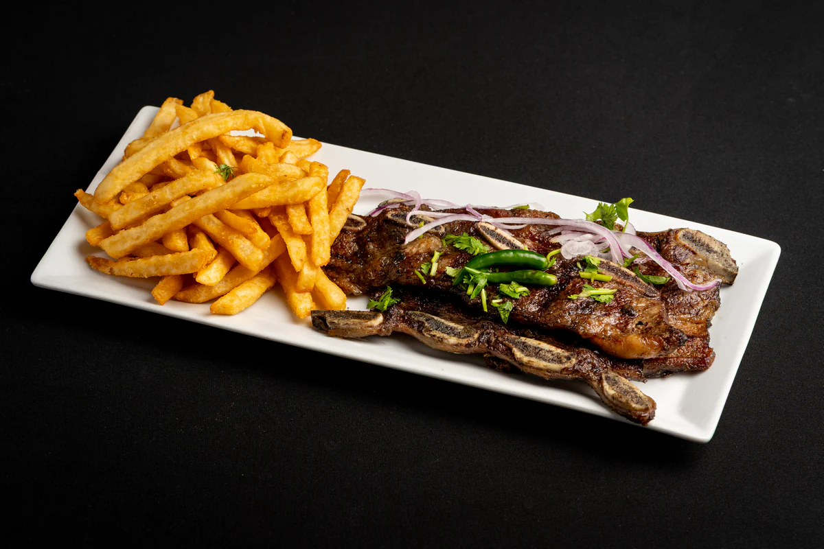 Grilled short ribs on a long white plate under sliced red onion and coriander, fries alongside.